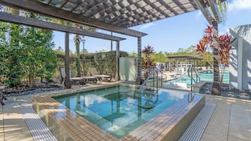 A swimming pool with a wooden pergola overhead, surrounded by tropical plants and a clear sky.