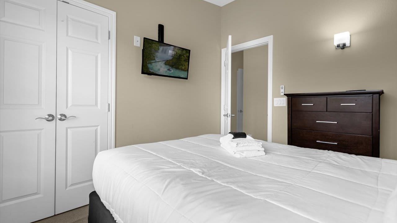 Un dormitorio con una cama, televisión montada en la pared, cómoda y una puerta.