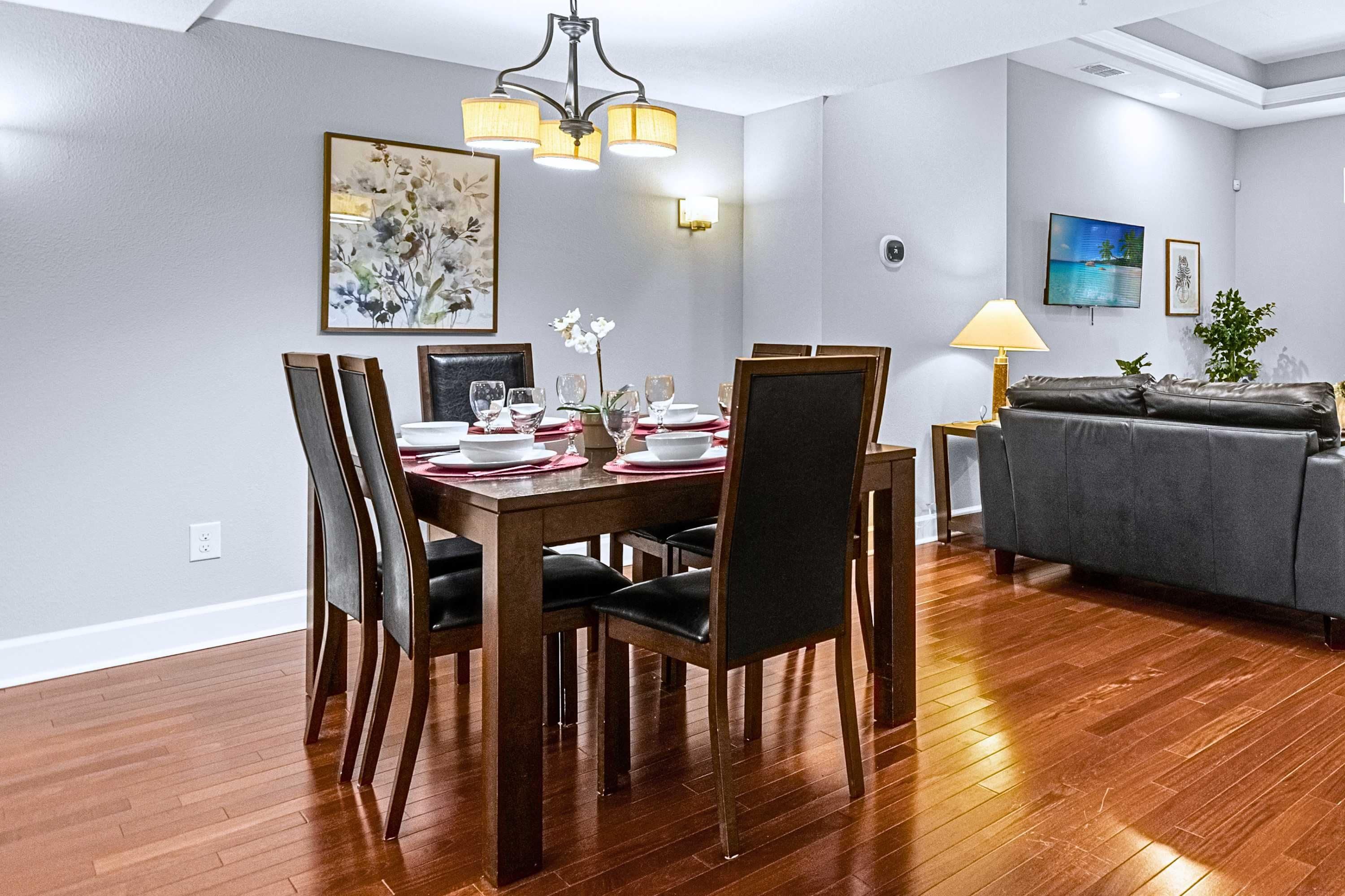 Uma sala de jantar com mesa de madeira posta para quatro pessoas, ladeada por cadeiras, um candelabro e um quadro na parede.