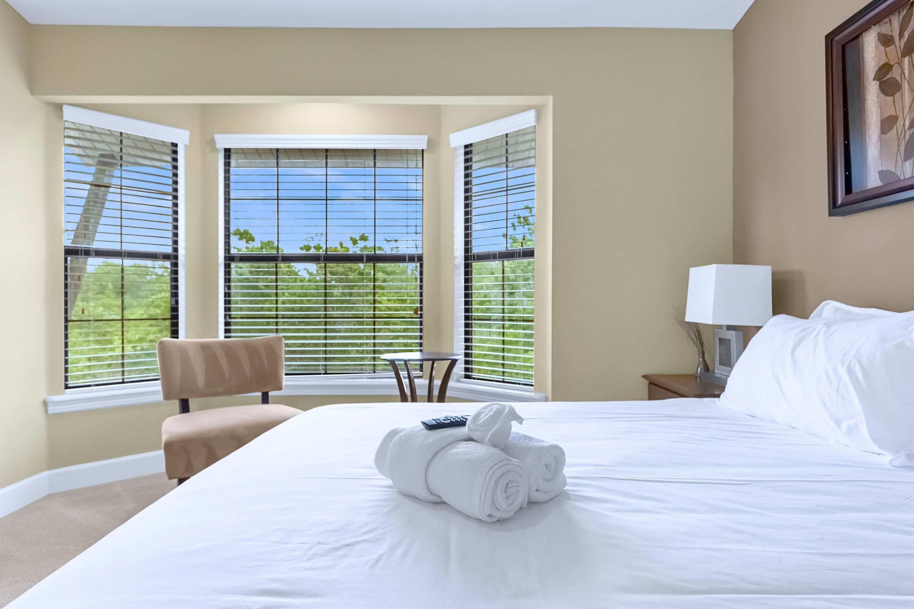 Quarto de hotel com cama grande, uma cadeira, mesa pequena e janela com persianas.