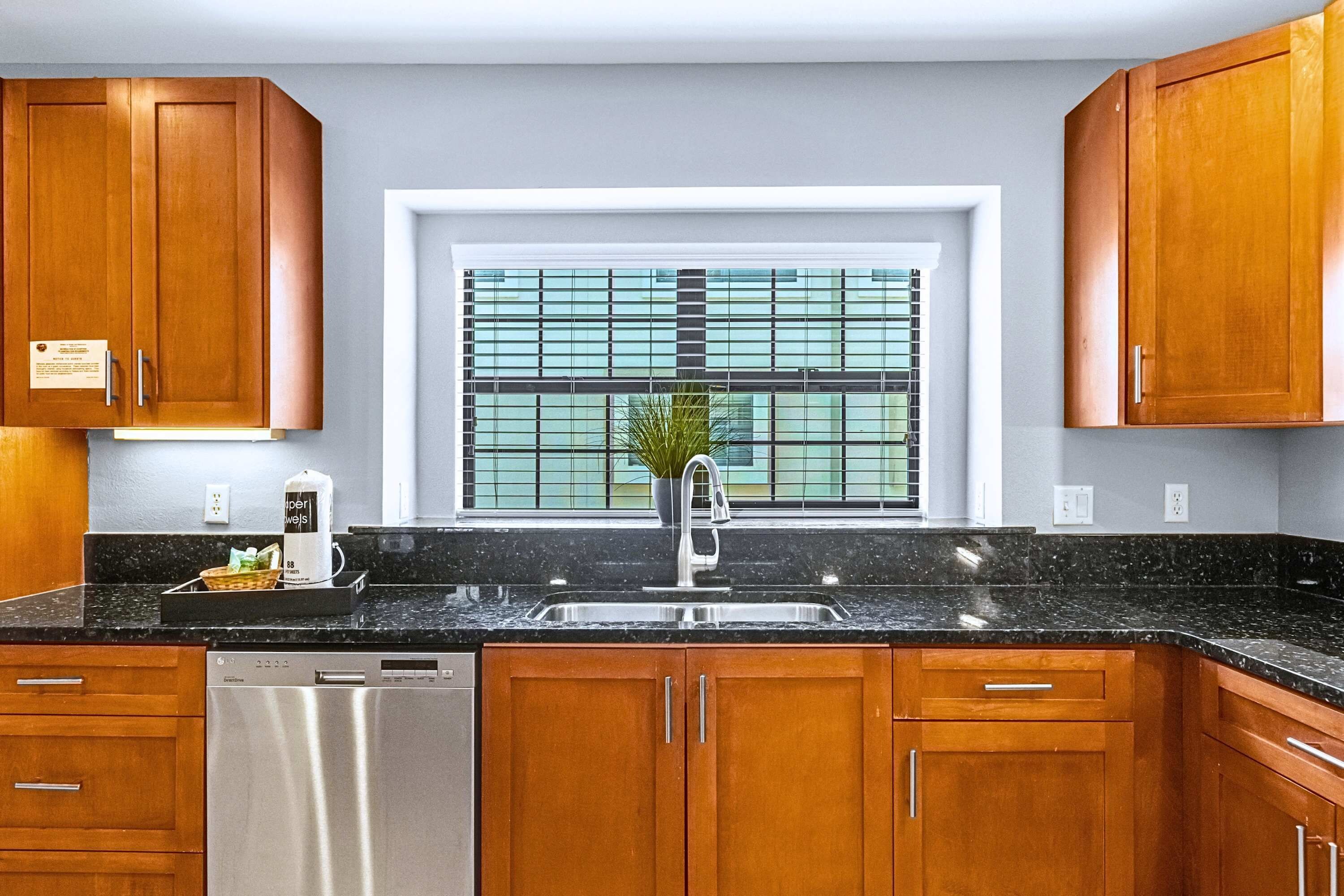 A kitchen with wooden cabinets, a black countertop, a stainless steel sink, and a window with blinds.