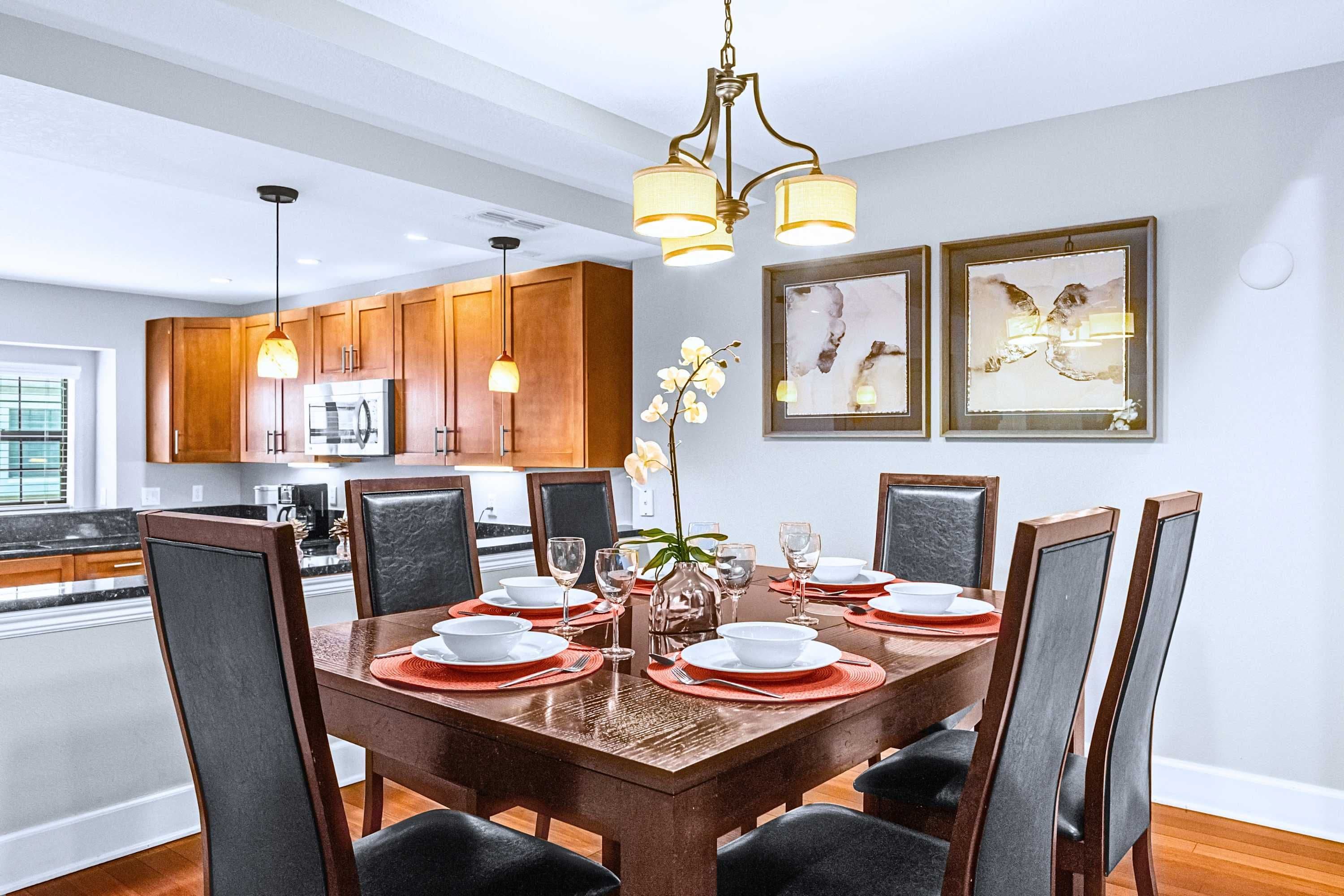 A dining area with a wooden table set for six, surrounded by chairs, in a modern kitchen with wooden cabinets and pendant lighting.