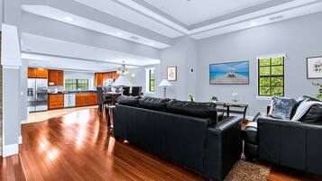 Ampla sala de estar com sofá seccional preto, mesa de jantar com cadeiras e cozinha ao fundo.
