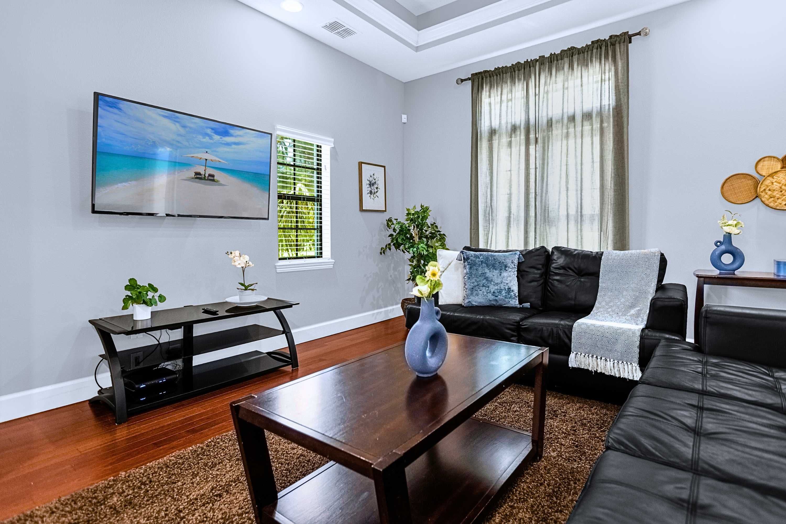 A modern living room with a flat-screen TV, a black leather sofa, a wooden coffee table, and a large window with curtains.