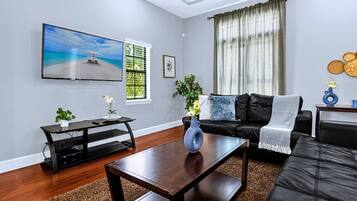 A modern living room with a flat-screen TV, a black leather sofa, a wooden coffee table, and a large window with curtains.