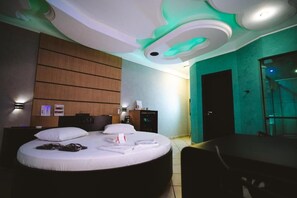 A hotel room with a round bed, a desk, and a chair. There is a wooden panel wall and a glass door leading to a balcony.