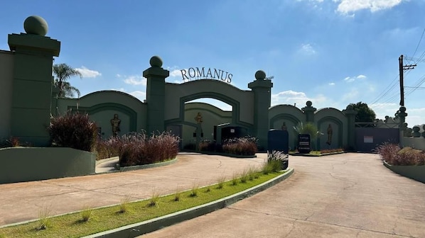 Une entrée fermée par un portail, sur laquelle on peut lire le nom « ROMANUS », le tout entouré de verdure et surplombé par un ciel dégagé.