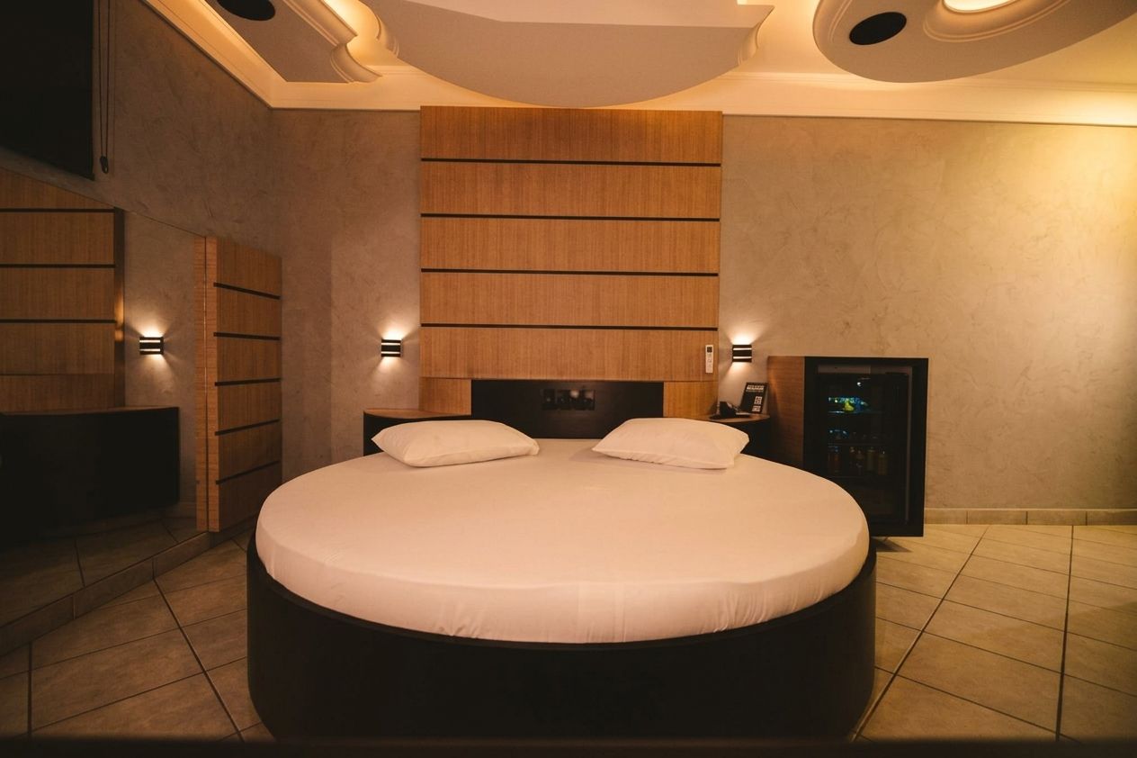 Un dormitorio moderno con una cama redonda grande, cabecera de madera integrada, iluminación en la pared y un minibar.
