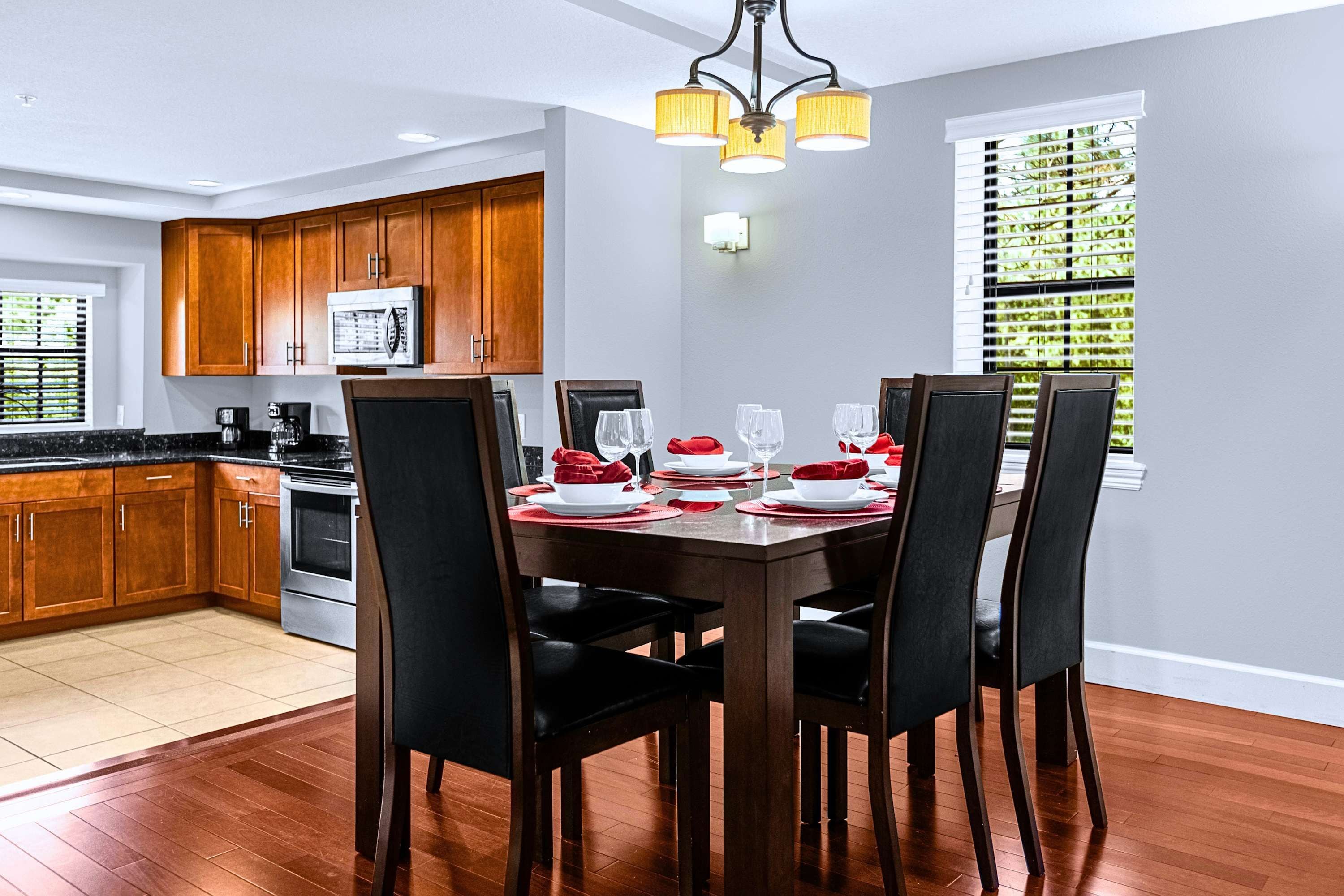 Comedor con mesa de madera dispuesta para cuatro personas, rodeada de sillas, en una cocina con armarios de madera y un microondas.