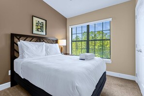 Une chambre d’hôtel avec un lit, une table de chevet, une lampe, une fenêtre avec des stores et un tableau encadré au mur.
