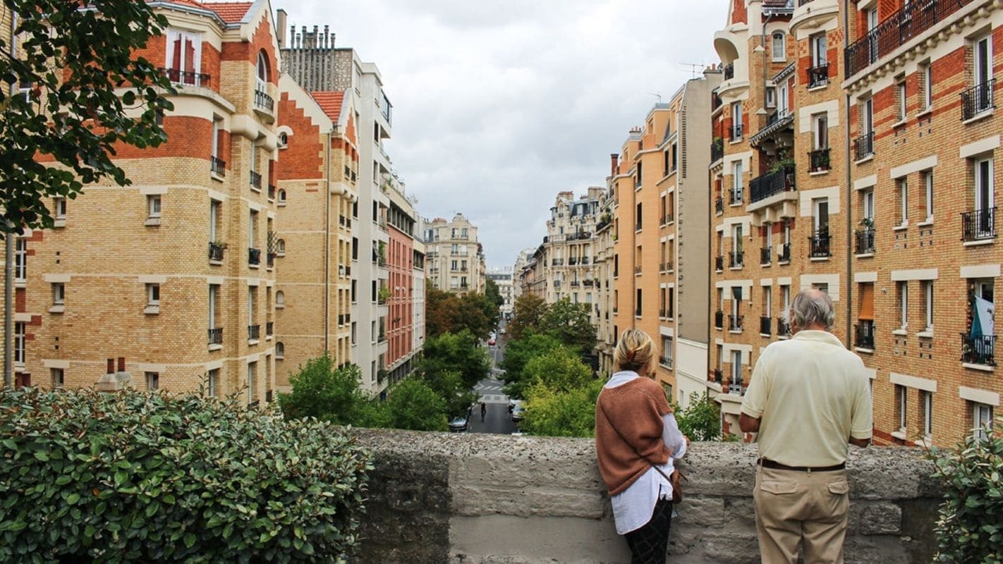 A narrow street flanked by multi-story buildings with balconies, a stone wall in the foreground, and a clear sky.