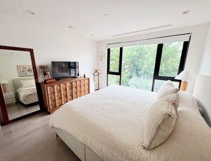 Een slaapkamer met een groot bed, een televisie op een houten kast, een spiegel en een raam met uitzicht op bomen.