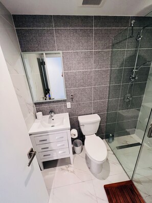 Een badkamer met een wit wastafelmeubel, spiegel en toilet, een douchegedeelte en een betegelde vloer.