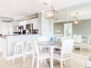 A modern kitchen with white cabinetry, a central island, and a dining area with white chairs and a round table. - ☀️Bay Views | Close to Beach | Golf Cart (LSV) | Harbour Pointe 806☀️ (Miramar Beach)