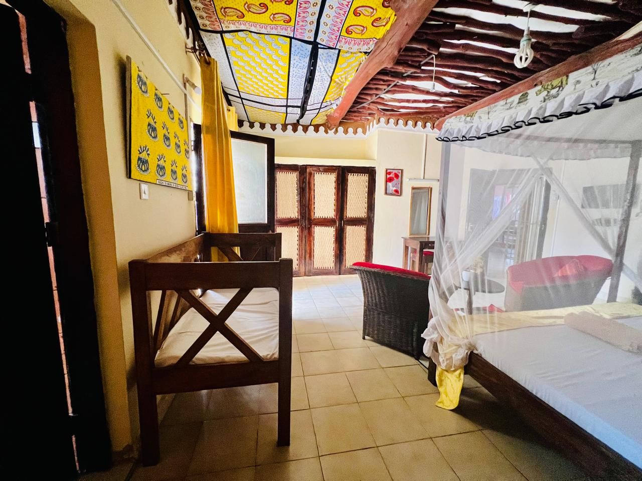 A room with a bed, a table, a chair, a wicker chair, a mosquito net, and a door with a wooden frame.