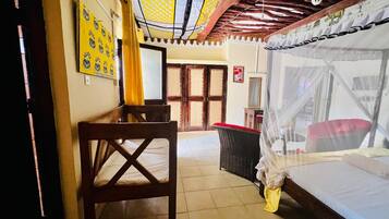 A room with a bed, a table, a chair, a wicker chair, a mosquito net, and a door with a wooden frame.