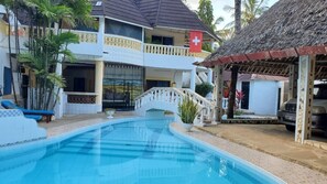 Un área de piscina con un puente, un edificio con balcón y una estructura con techo de paja.