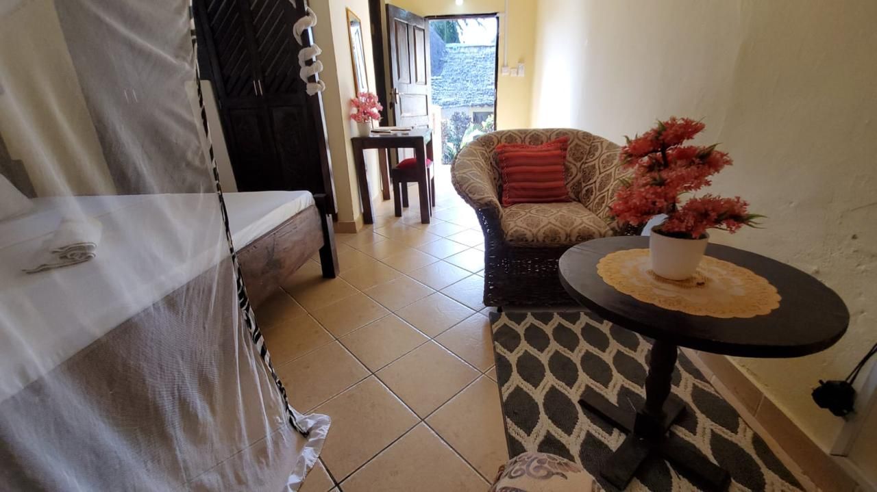 A room with a bed, a sofa, a small table with a plant, and a dining area in the background.