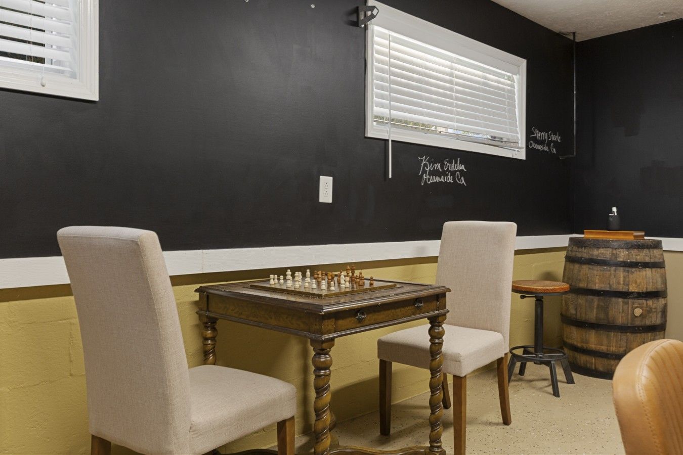 Una habitación con una mesa de ajedrez, dos sillas, un barril de madera y una ventana con persianas.