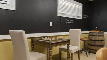 Una habitación con una mesa de ajedrez, dos sillas, un barril de madera y una ventana con persianas.