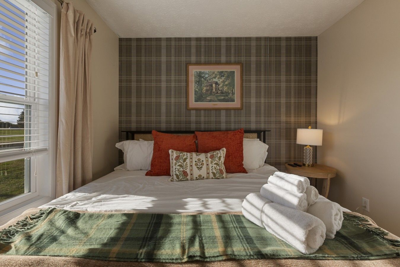 A hotel room with a bed, a nightstand with a lamp, a window with curtains, and a plaid-patterned wall.