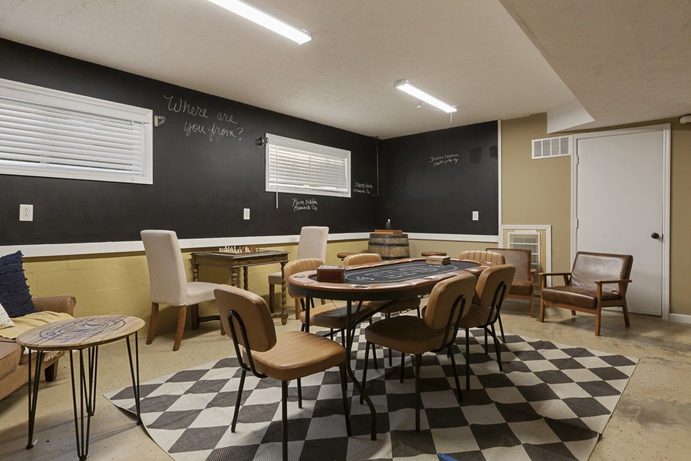Una habitación con piso a cuadros, una mesa redonda y varias sillas. Hay dos ventanas con persianas, una puerta y una pizarra en la pared.