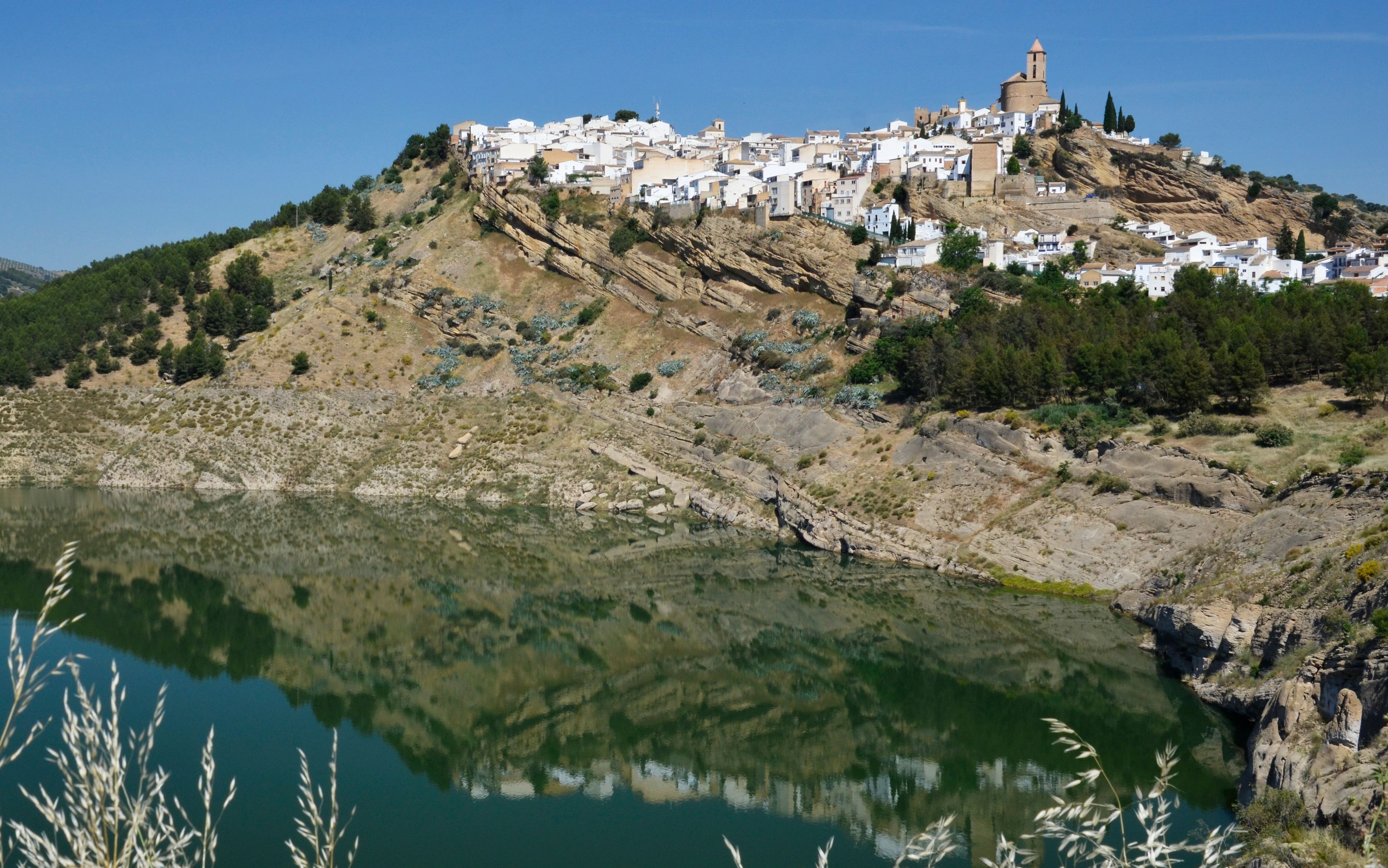 Un pueblo costero con edificios blancos en una colina, una masa de agua en primer plano y un cielo despejado.
