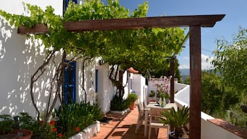 Un edificio blanco con contraventanas azules, una pérgola de madera, plantas en macetas y un patio con mesa y sillas.