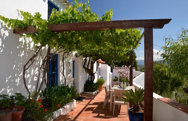 Edifici blanc amb persianes blaves, una pèrgola de fusta, plantes en testos i un patí amb taula i cadires.