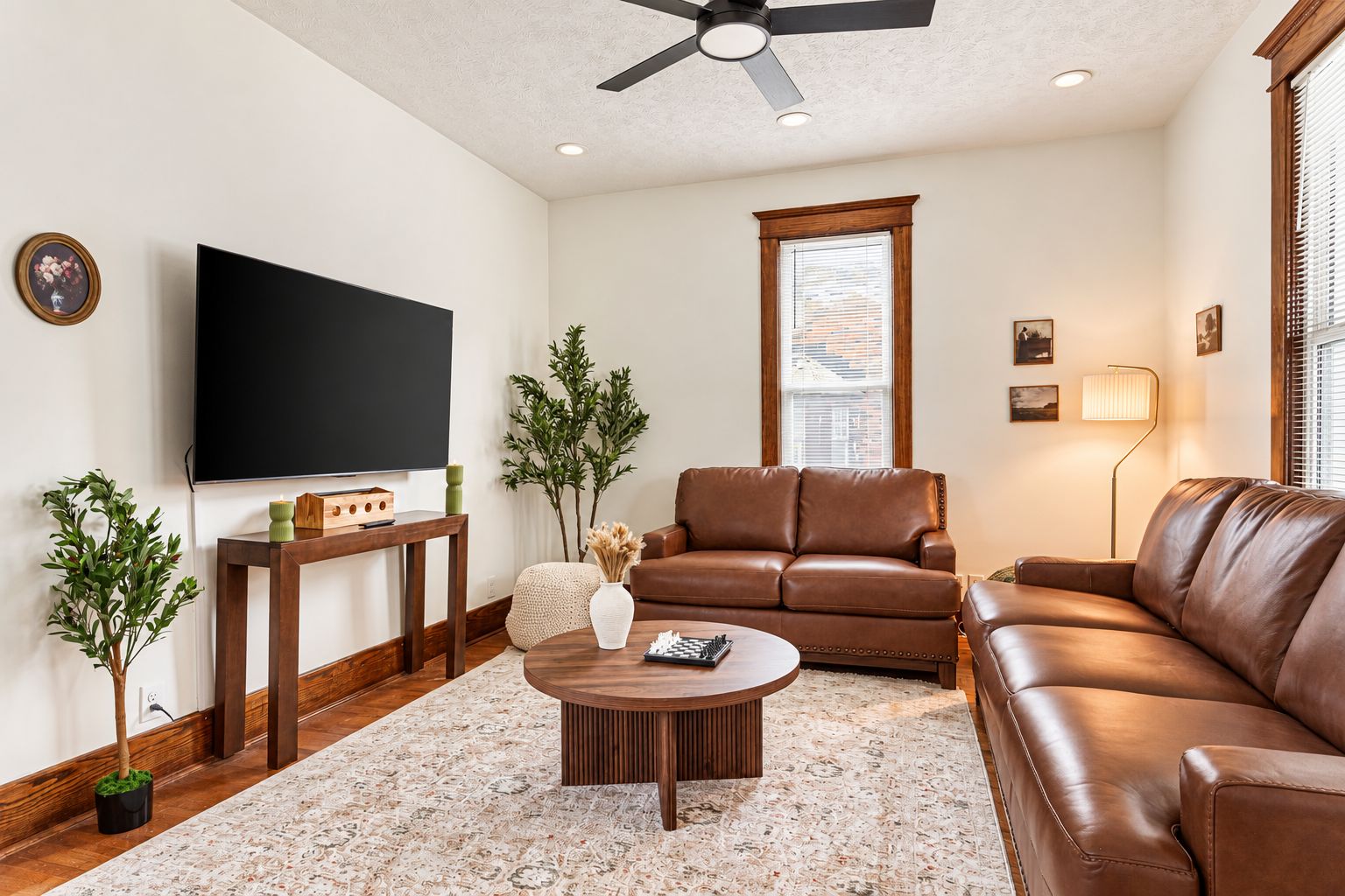 A living room with a flat-screen TV, leather sofas, a coffee table, and a large window with blinds.