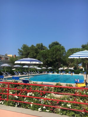 A swimming pool with lounge chairs and umbrellas, surrounded by greenery and a red railing. - Holiday Home 'Bilocale Con Terrazza E Piscina' with Air Conditioning (Marina di Bibbona)