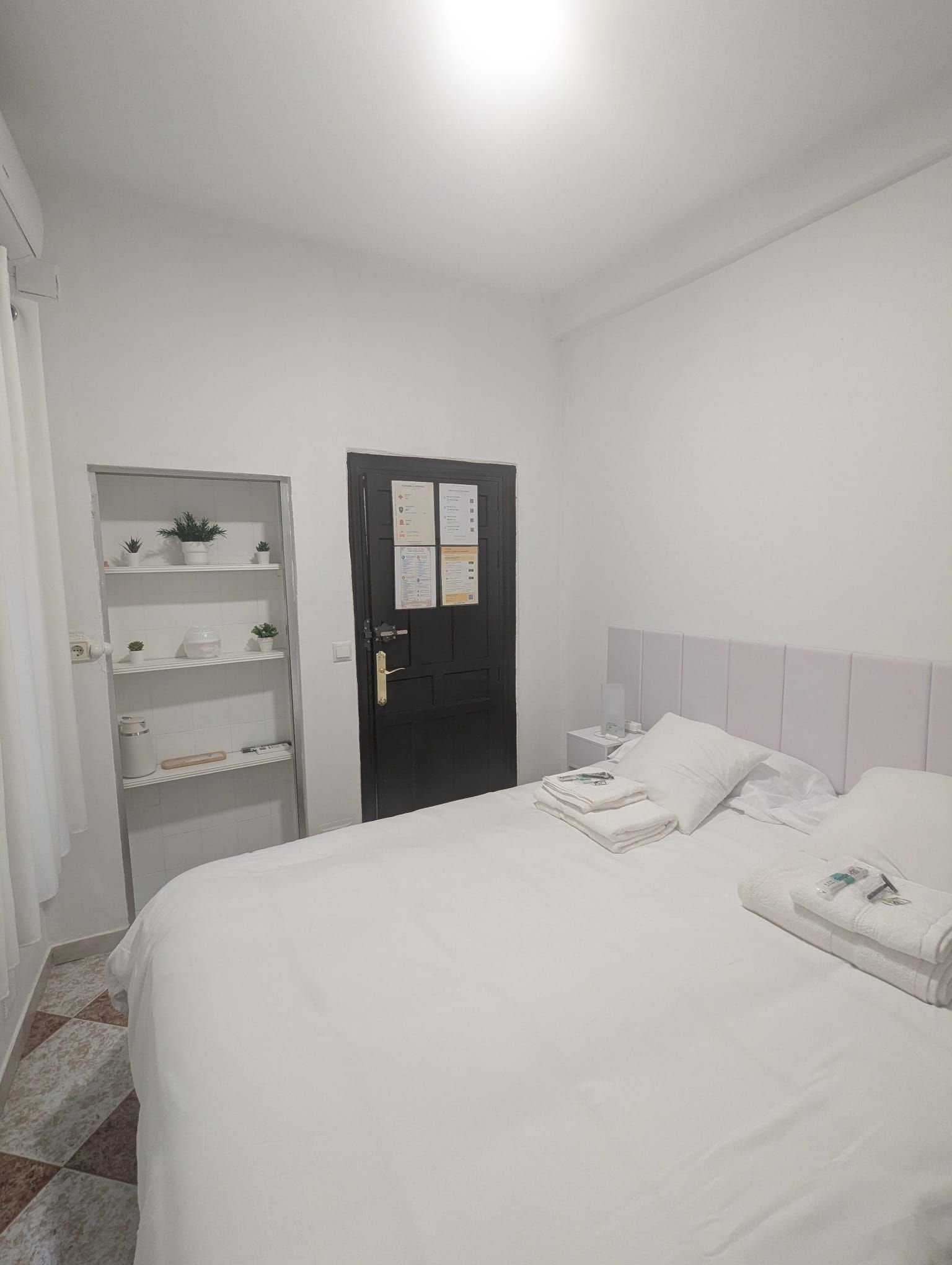 Hotellihuone, jossa on sänky, vaatekaappi, ilmoitustaululla varustettu ovi ja yöpöytä, jossa on lamppu.