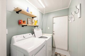 A laundry area with a front-loading washer and dryer, a shelf with cleaning supplies, and a door with hooks. - Free Heated Pool Home, Luxury Beds, Pet Friendly (Cape Coral)