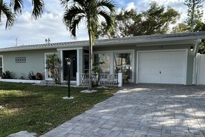 Ein Bungalow mit Garage, gepflasterter Auffahrt und Garten mit kleinen Sträuchern und einer Palme.