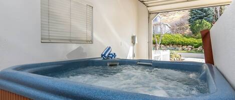 A hot tub with a wooden bench and a towel, a window with blinds, and a view of a garden outside.