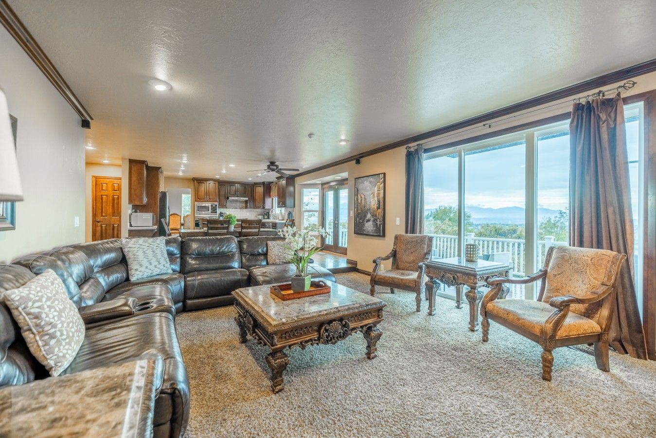 A spacious living room with a leather sofa, armchairs, and a coffee table. There is a large window with a view of the outdoors.