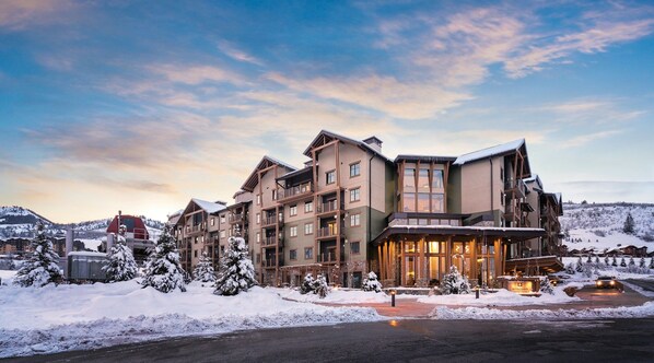 Ein mehrstöckiges Hotel vor der Kulisse eines Skigebiets, mit einem markanten Eingangsbereich und einer beleuchteten Fassade, eingebettet in eine verschneite Landschaft.