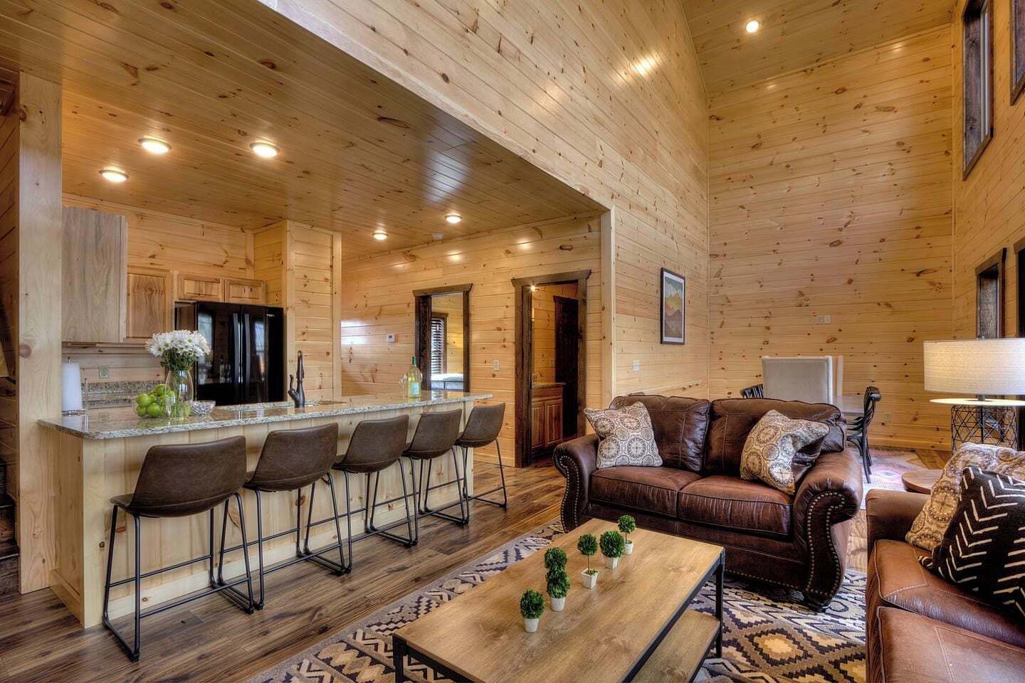 A spacious living area with a wooden interior, a bar with stools, a leather sofa, and a coffee table with potted plants.