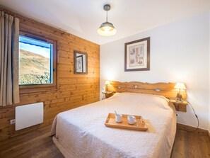Et soveværelse med sengegavl i træ, et stort vindue med gardiner, et natbord med lampe og en bakke med kopper og ske.