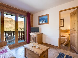 A room with a wooden cabinet, a television, a bed, and a view of a mountainous landscape through a sliding door. - Bright apartment near the slopes (Les Menuires)