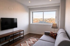 Uma sala de estar com televisão de tela plana, um móvel de madeira para televisão, um sofá com almofada colorida, um abajur e uma grande janela com vista para a cidade.