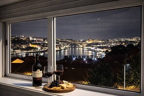 A bottle of wine, two glasses, and a cheese board on a windowsill with a view of a cityscape at night. - Panoramic Douro View | Bridge & City Centre (Vila Nova de Gaia)