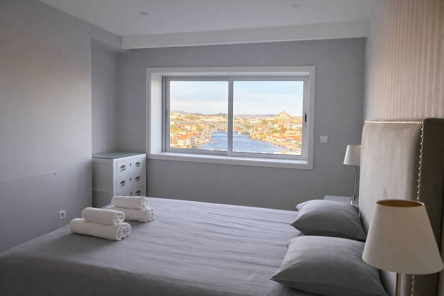 Une chambre d’hôtel dotée d’une grande fenêtre, d’un lit, d’une table de chevet avec des lampes et offrant une vue sur le paysage urbain.