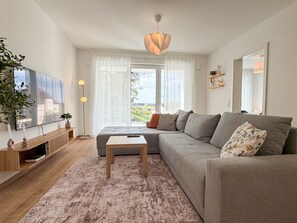 A modern living room with a large flat-screen TV, a grey sofa, a small wooden coffee table, and a large window with a view of greenery.
