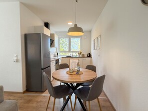 A modern kitchen with a dining area, featuring a round wooden table with chairs, a hanging light fixture, and a window with a view of greenery.
