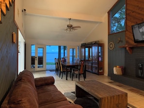 A spacious living area with a wooden coffee table, a brown sofa, and a dining table with chairs. There is a fireplace with a TV mounted above it, and a large window offering a view of the outdoors. - Waterfront Spacious 4-bedroom house on Toledo Bend in Hemphill (Hemphill)