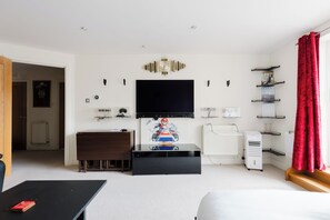 A living room with a flat-screen TV, a gaming console, a radiator, and a window with red curtains. - Studio Flat with Waterfront Views in East London (Greater London)