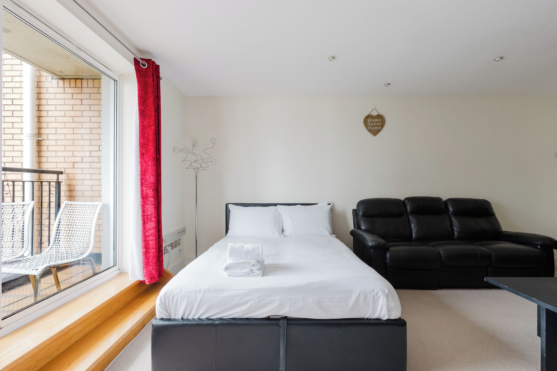 A hotel room with a bed, a black leather sofa, a small table, and a window with a red curtain.