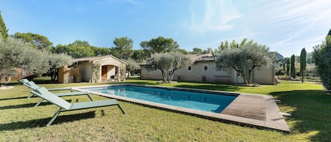 Una piscina con sillas de descanso, rodeada de un césped cuidado y árboles, con una casa al fondo.