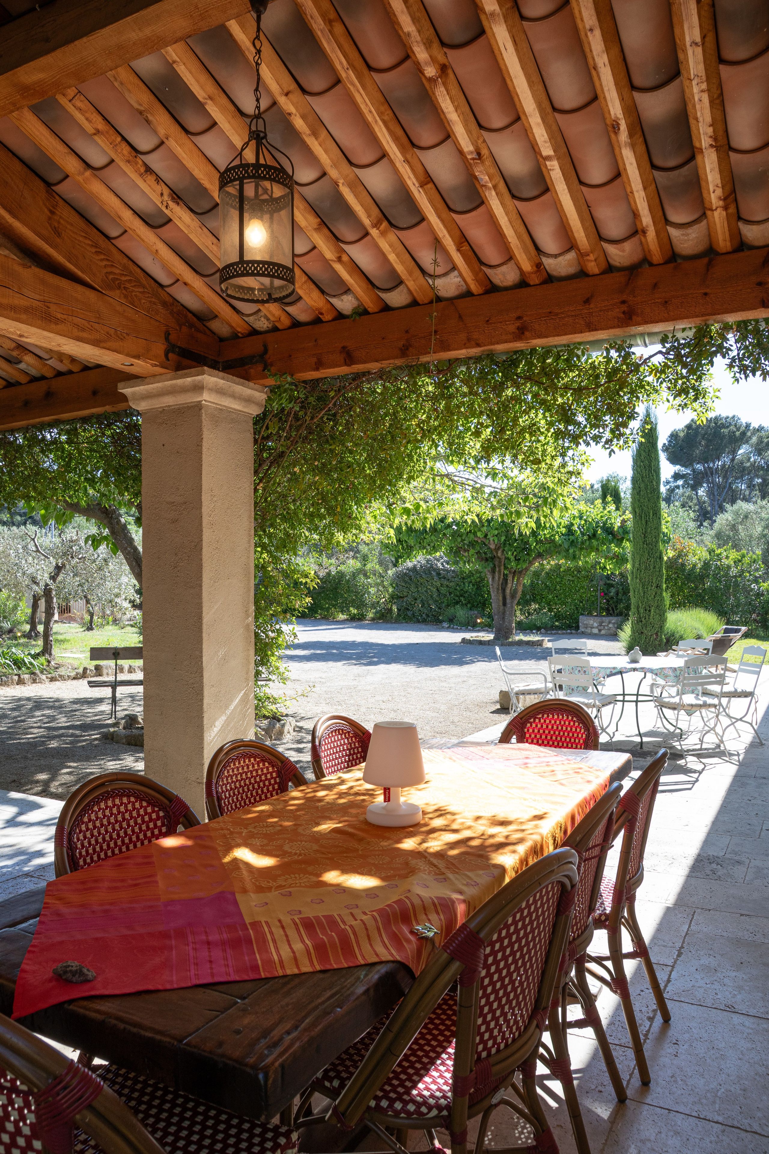 Une terrasse extérieure dotée d’une pergola en bois, d’une table entourée de chaises et offrant une vue sur un jardin avec des arbres et un chemin de gravier.